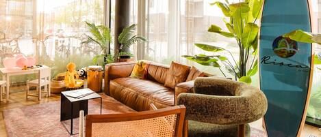 A modern living room with a leather sofa, a wooden chair, a surfboard, and potted plants.