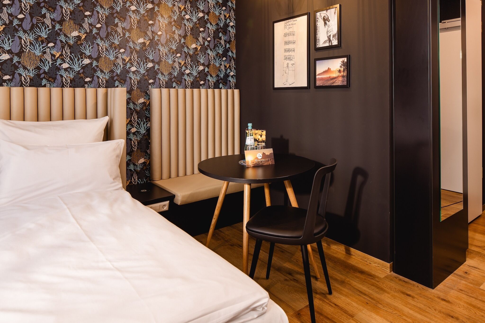 Habitación de hotel con cama, una mesa redonda con una silla, una mesita con una botella y un menú, una pared decorada con cuadros, y suelo de madera.