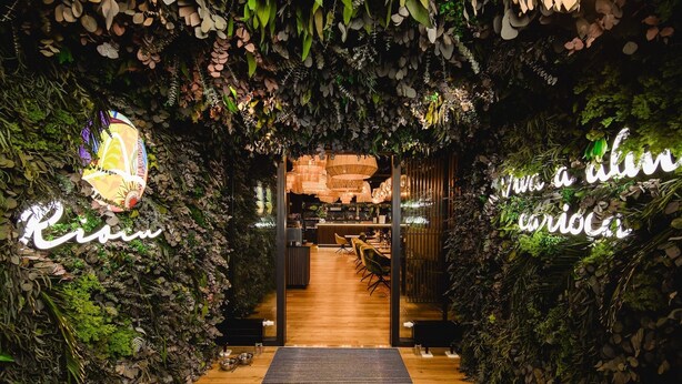 An indoor pathway leading to a dining area with a wooden floor and a neon sign reading "Café". - Rioca Konstanz Posto 10 (Konstanz)