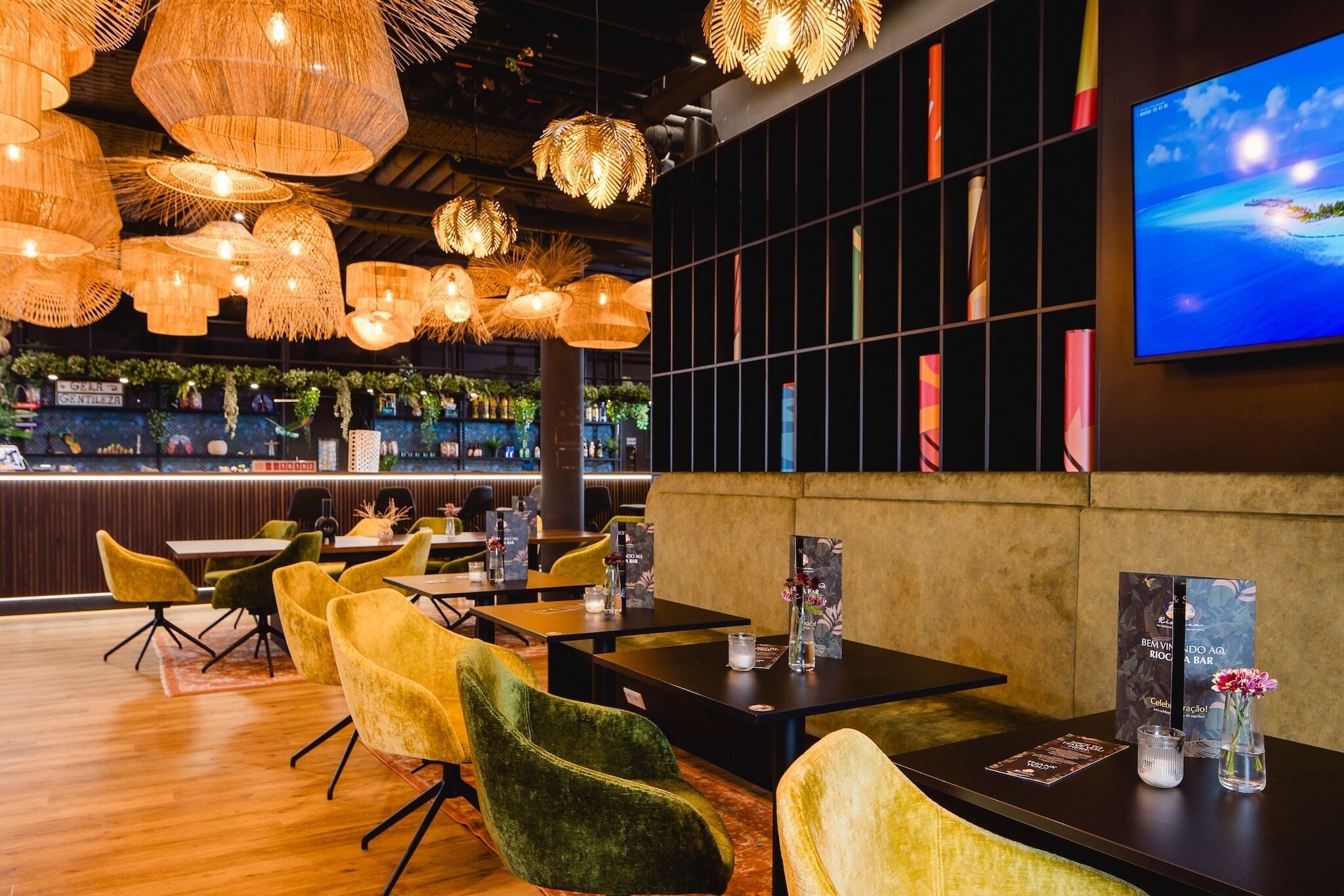 Een restaurant met rieten plafondlampen, groene fluweelstoelen en een bar met een televisiescherm.