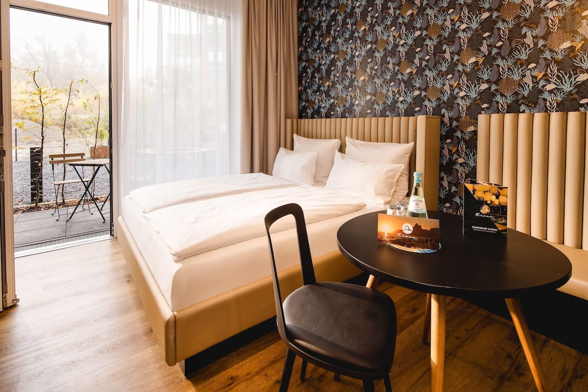 A hotel room with a bed, a dining table, a chair, and a view of the outdoors.