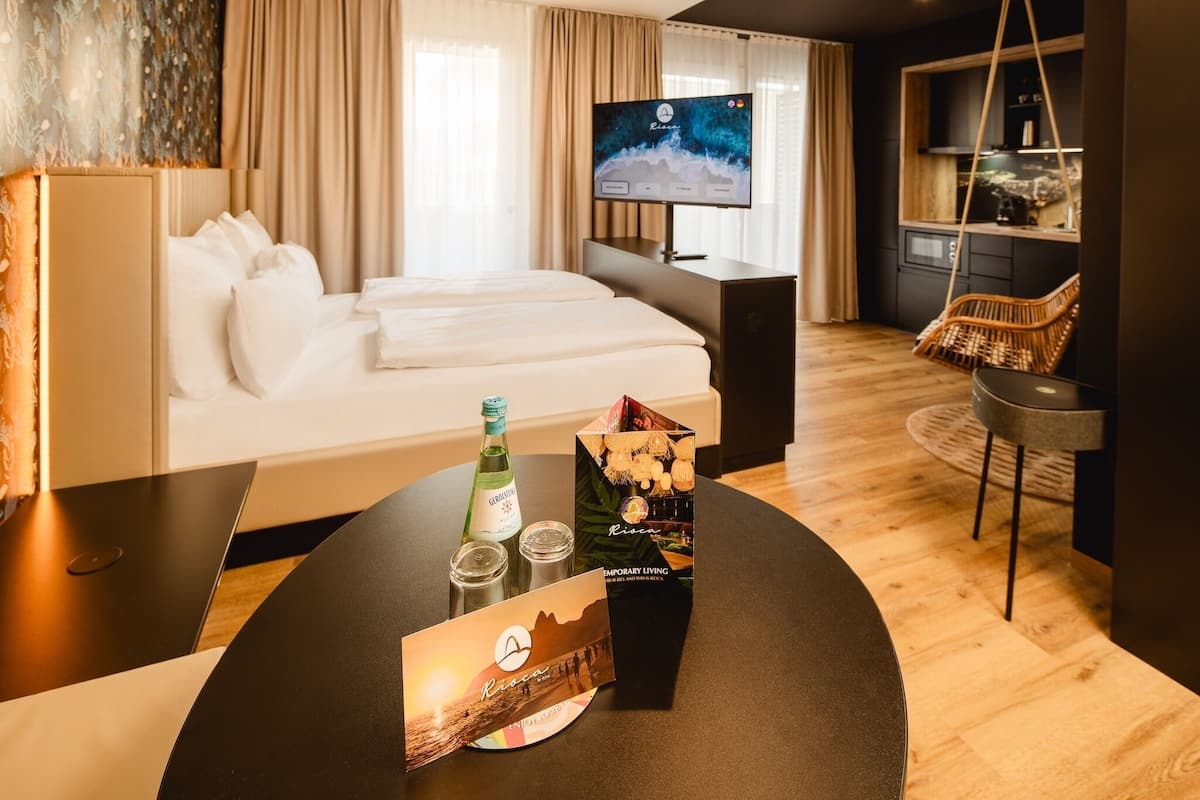 A hotel room with a bed, a desk, a television, and a dining table with a bottle and glasses.