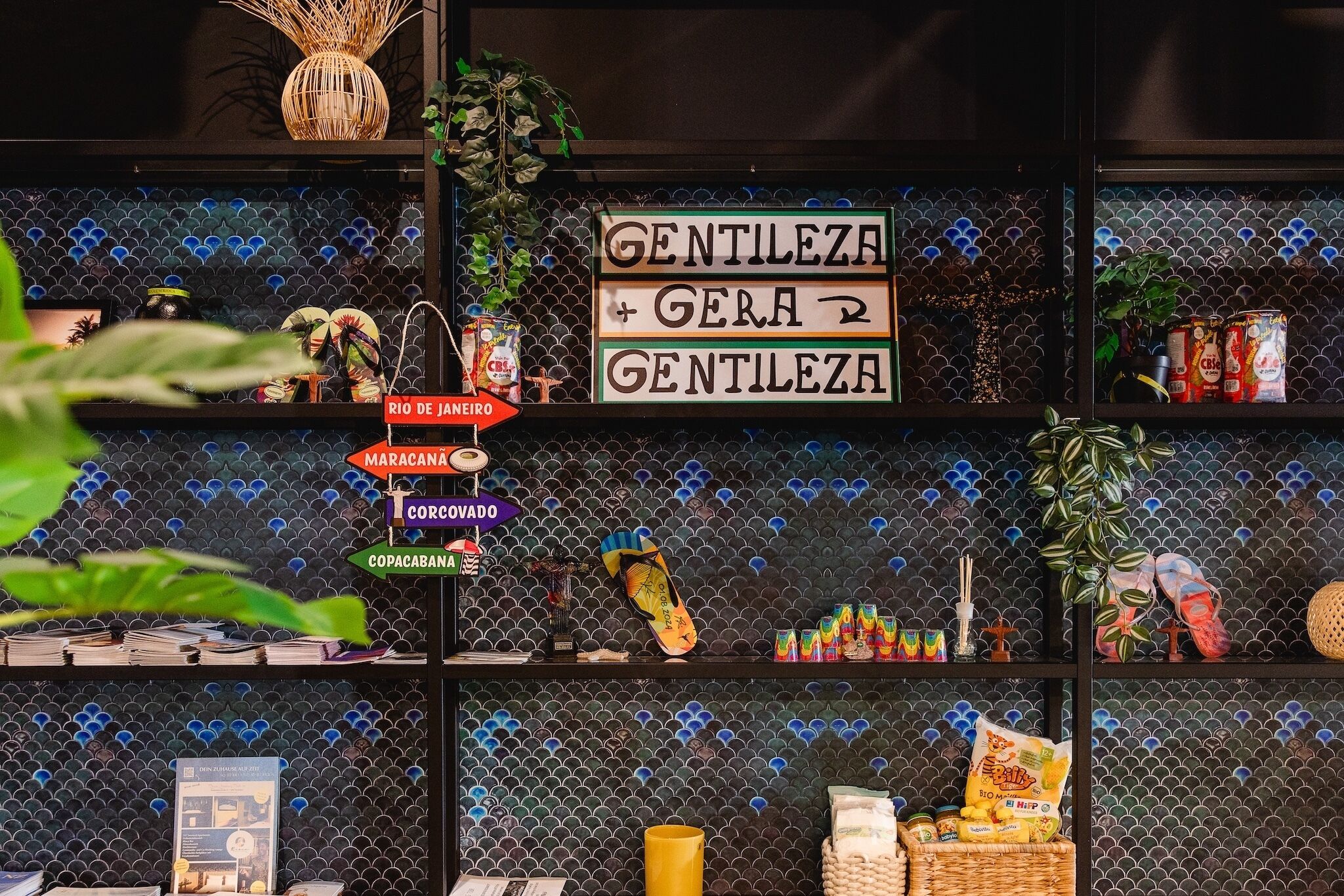 Une pièce avec un mur recouvert de carreaux, une pancarte sur laquelle on peut lire « Gentileza + Gera + Gentileza », et diverses étagères contenant des collations et une plante.