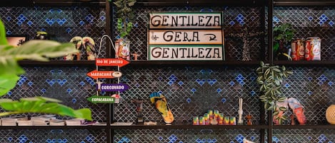 Una habitación con pared de azulejos, un cartel que dice 'Gentileza + Gera + Gentileza' y varios objetos en estanterías, como aperitivos y una planta.