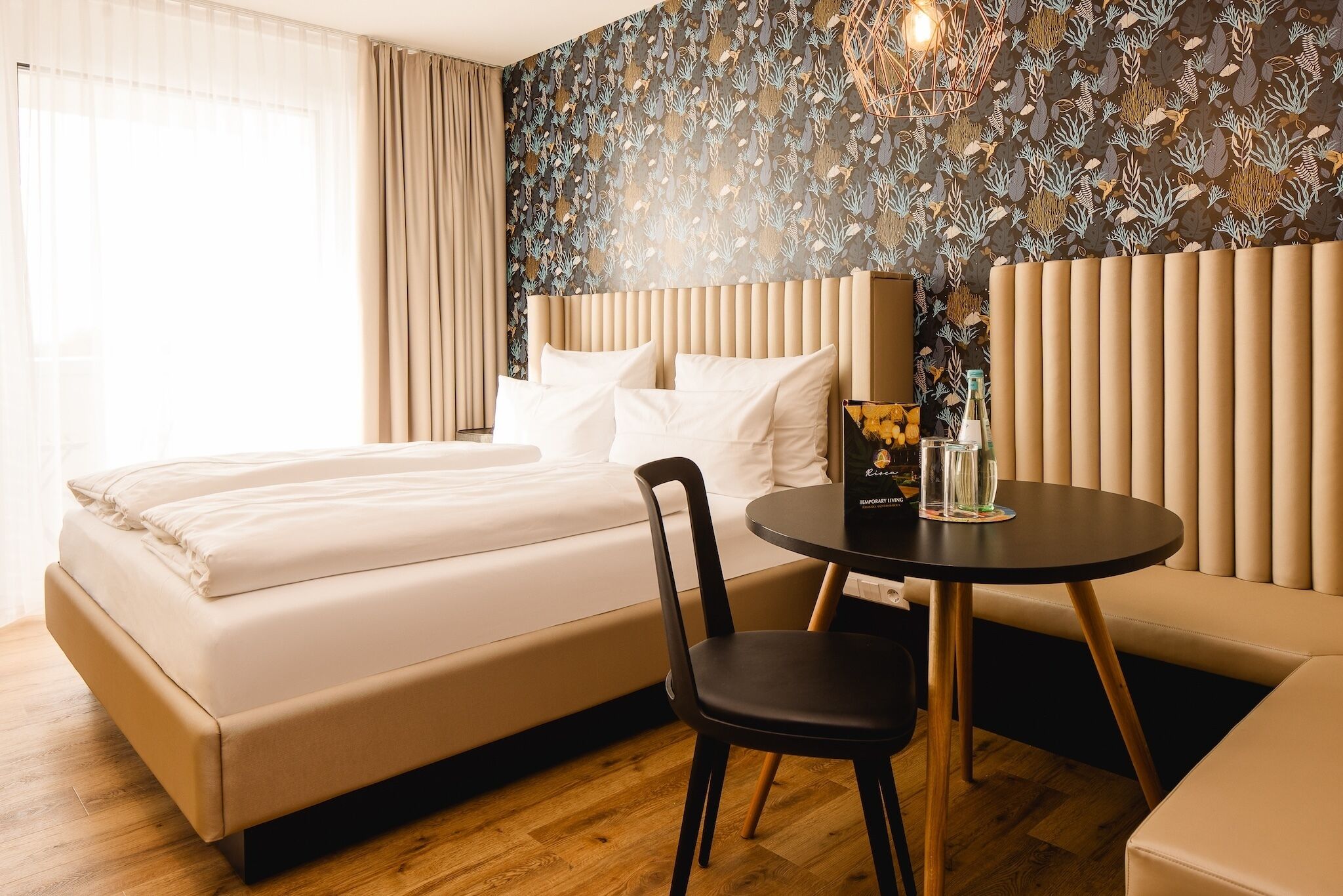 Une chambre d’hôtel avec un lit, une chaise, une table ronde et un mur orné d’un motif de feuilles.