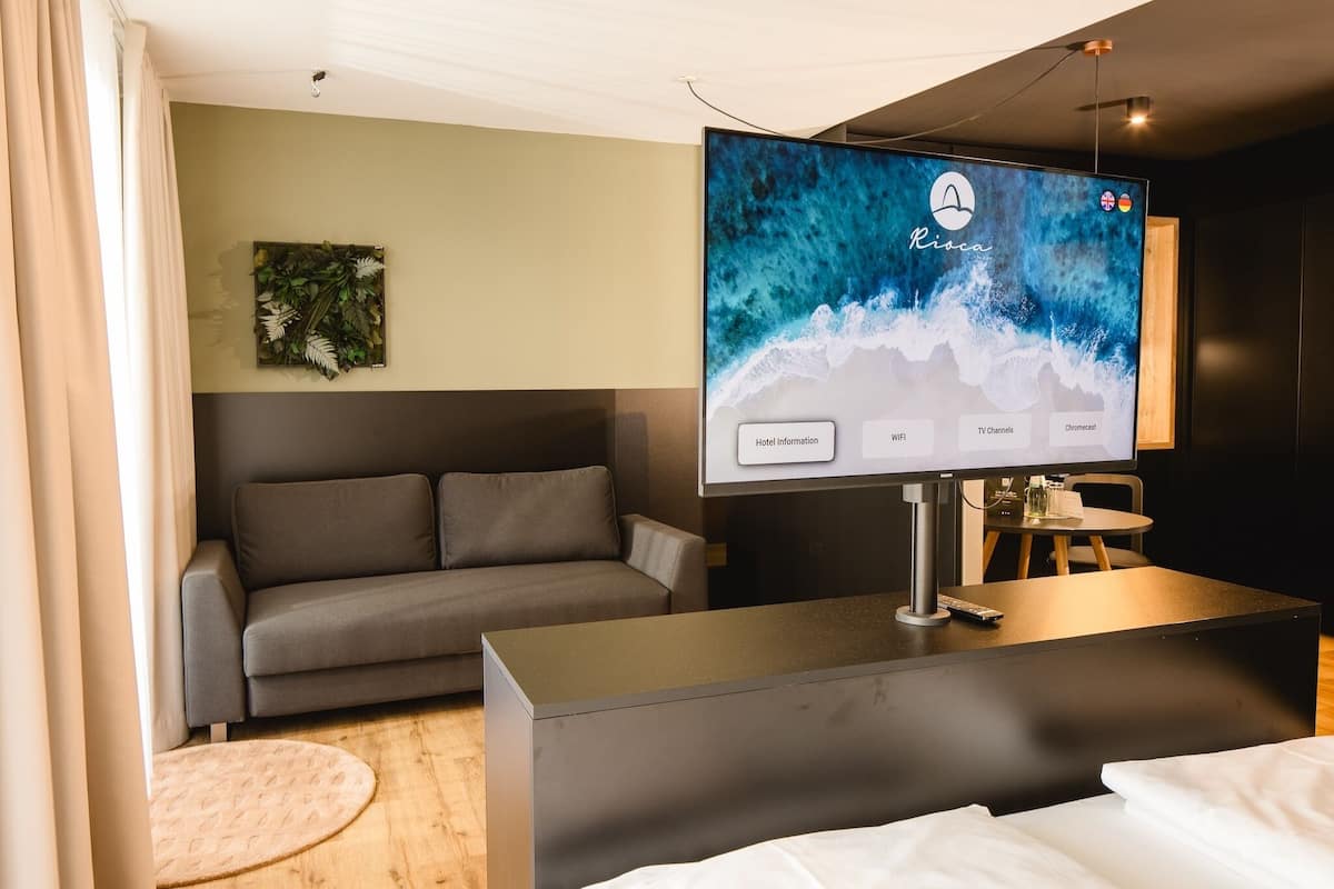 A modern hotel room with a large flat-screen TV displaying the Riva logo, a grey sofa, a small round table, and a bed with white linens.