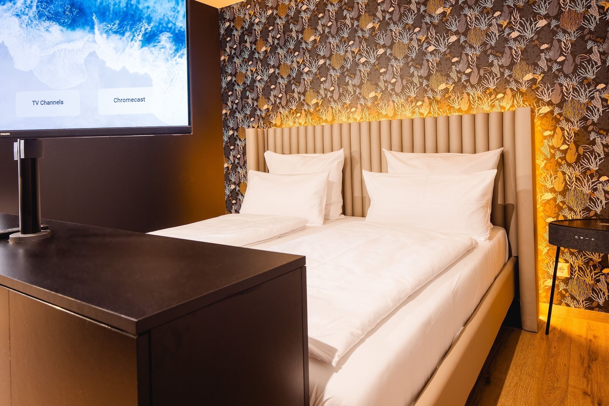 Una habitación de hotel con cama, televisor sobre un soporte y pared con diseño de hojas.
