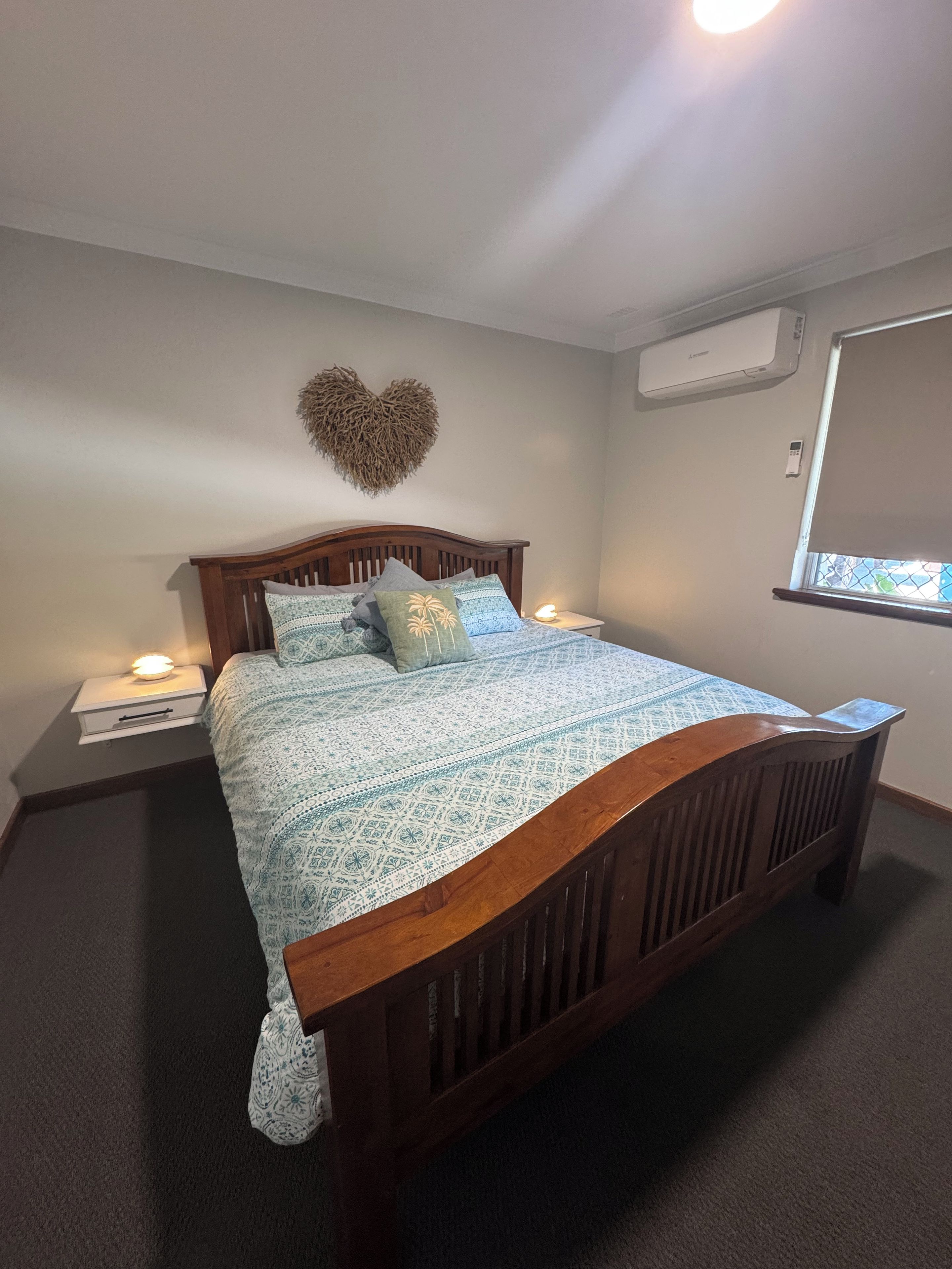 A bedroom with a wooden bed, patterned bedding, a heart-shaped wall decoration, bedside lamps, and an air conditioning unit.