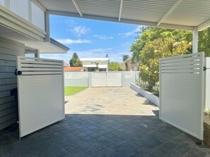 A covered outdoor area with a paved floor, two white gates, and a view of a residential building in the background.