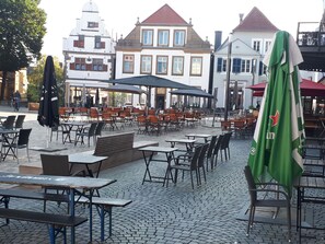Café seating area with tables and chairs on cobblestone pavement, offering shade under umbrellas.