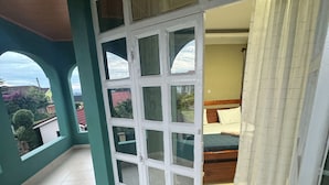 Una stanza con un'ampia porta-finestra scorrevole che dà su un balcone. La stanza presenta un letto con testiera in legno e una tenda alla finestra. Il balcone ha un pavimento piastrellato e si affaccia sui dintorni.