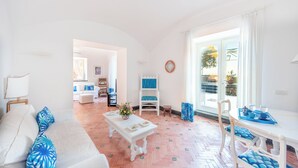 Un salon lumineux avec un canapé blanc, une table basse et un coin repas comprenant une table et des chaises blanches. Une grande fenêtre offre une vue sur l’extérieur.