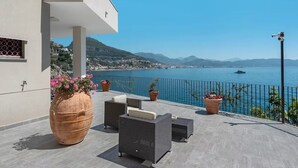 A terrace with outdoor furniture, a large vase with flowers, and a view of the sea and mountains.