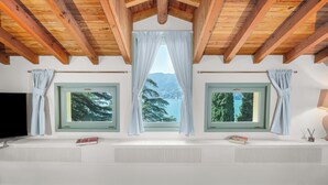 Une pièce avec un plafond en pente, des poutres en bois, deux fenêtres avec des rideaux, un radiateur et un téléviseur à écran plat.