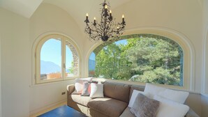Una habitación con un sofá beige, una mesa de centro oscura y una araña. Hay dos ventanas arqueadas amplias que ofrecen vistas a zonas verdes y montañas.