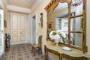 A hallway with a patterned floor, a mirror, a vase with flowers, and a chair.