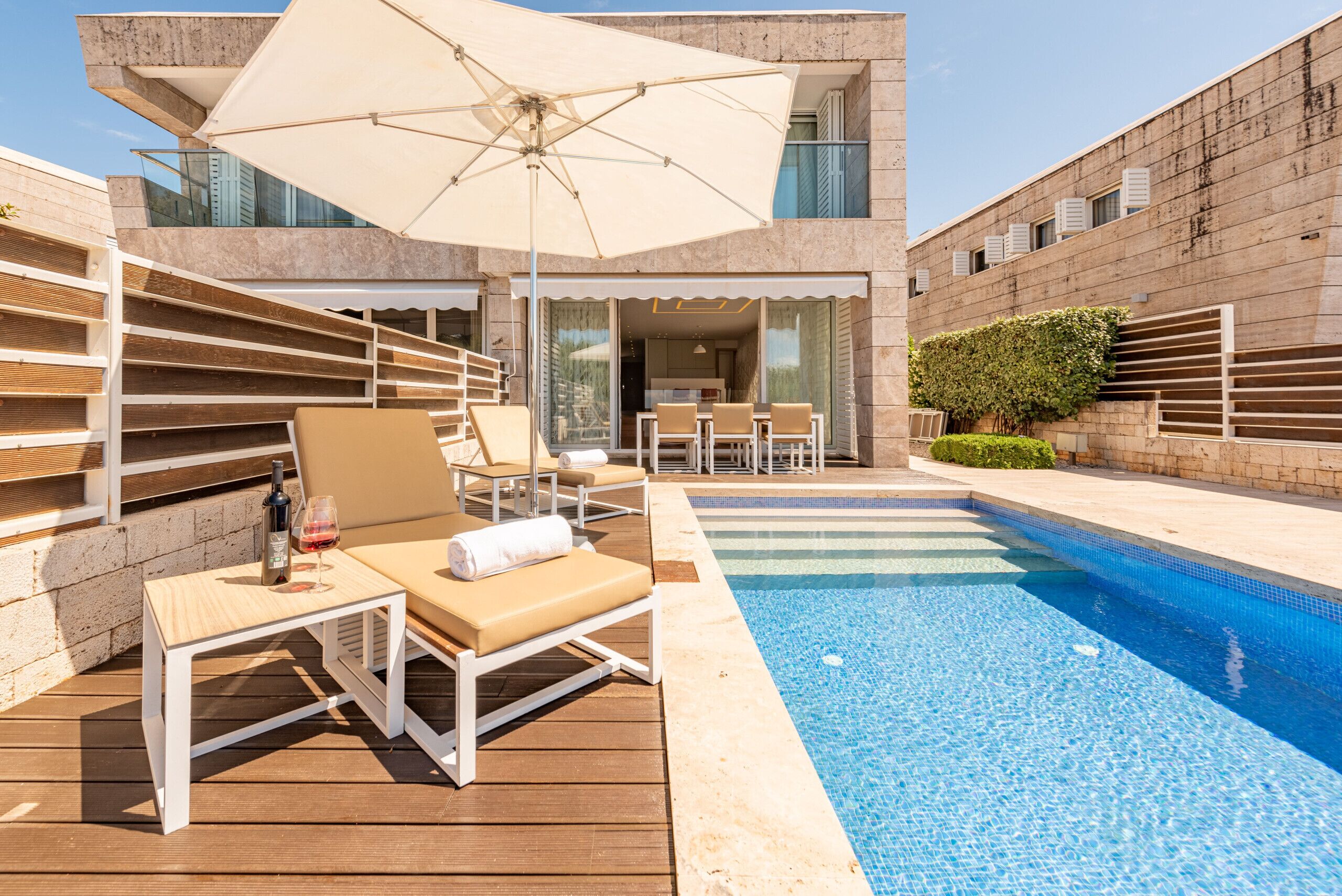 A modern outdoor pool area with lounge chairs, a table with a bottle and glass, and a large umbrella.