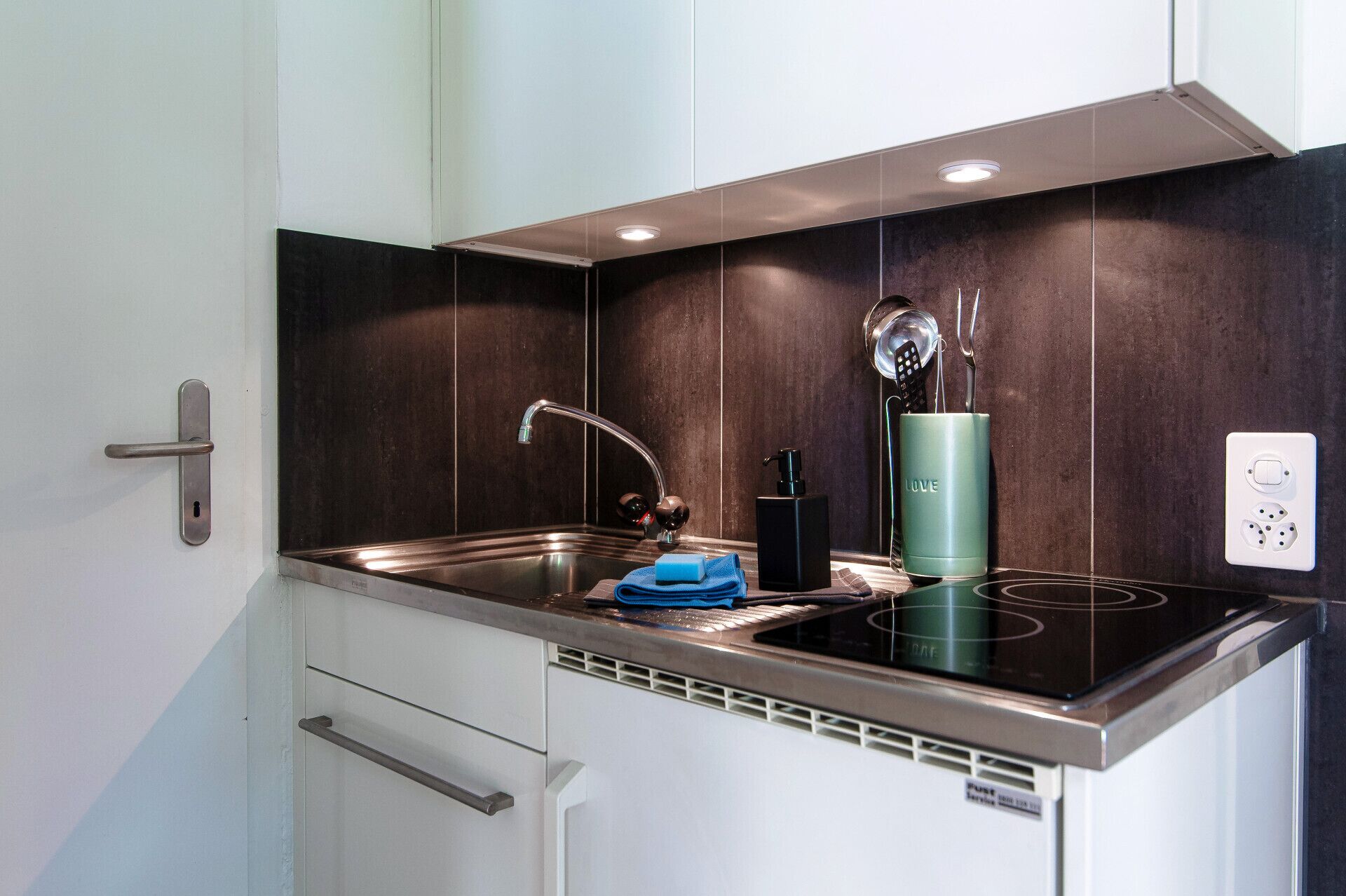 A compact kitchen with a stainless steel sink, a black faucet, a green tumbler, and a black soap dispenser.