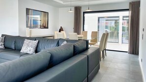 Una sala de estar moderna con un sofá seccional, un comedor con una mesa de vidrio y un amplio ventanal corredizo que da a un área exterior.