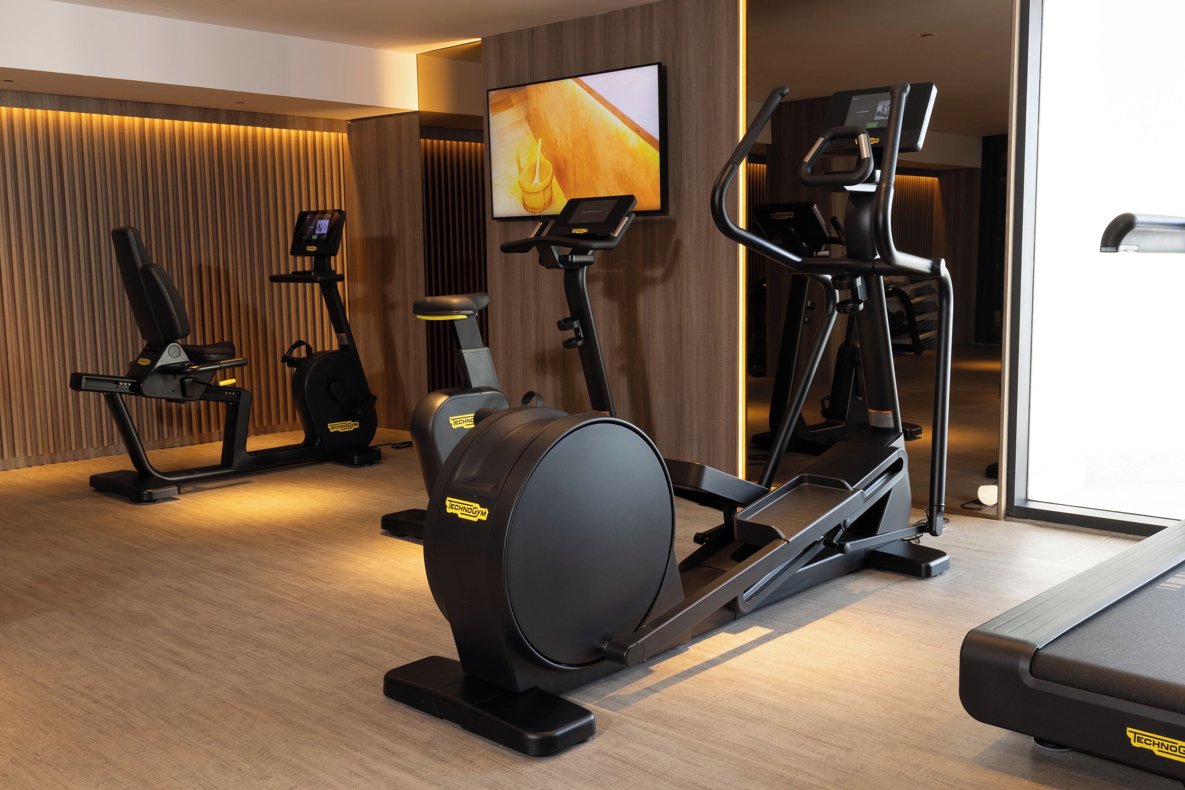 A modern gym with various exercise machines, a large TV screen, and a wooden wall design.