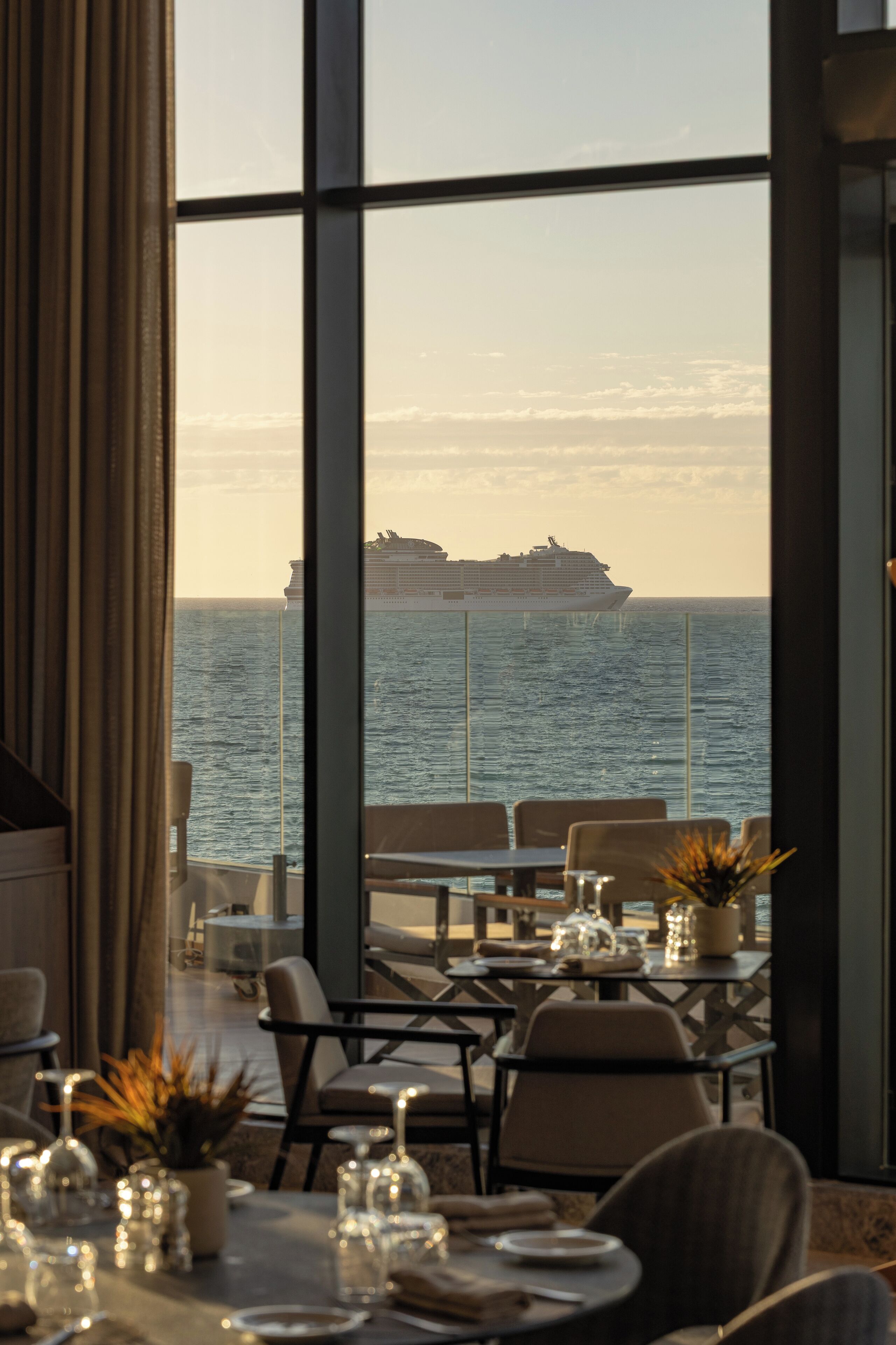 A dining area with tables set for a meal, large windows offering a view of the ocean and a cruise ship, and a potted plant on the table.