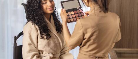 A person seated in a chair with a makeup palette in hand, another person assisting with makeup application.