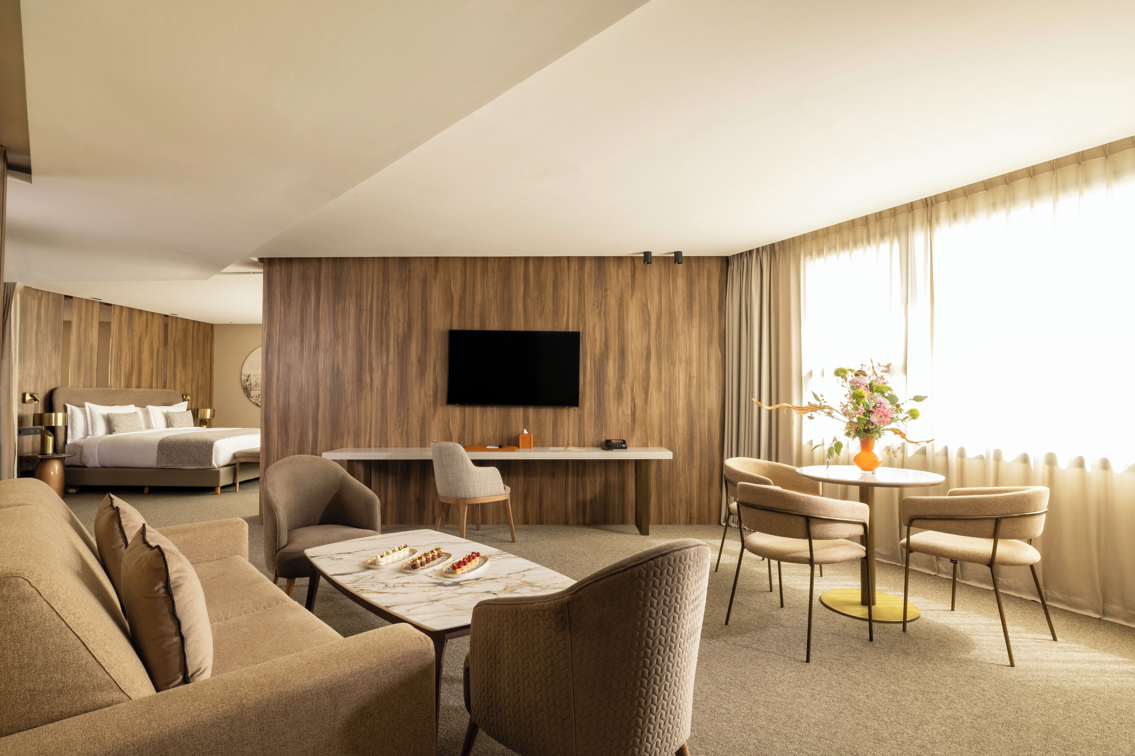 A hotel room with a bed, a desk, a dining table with chairs, a sofa, and a TV mounted on the wall.