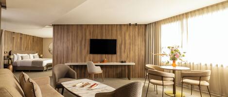 A hotel room with a bed, a desk, a dining table with chairs, a sofa, and a TV mounted on the wall.