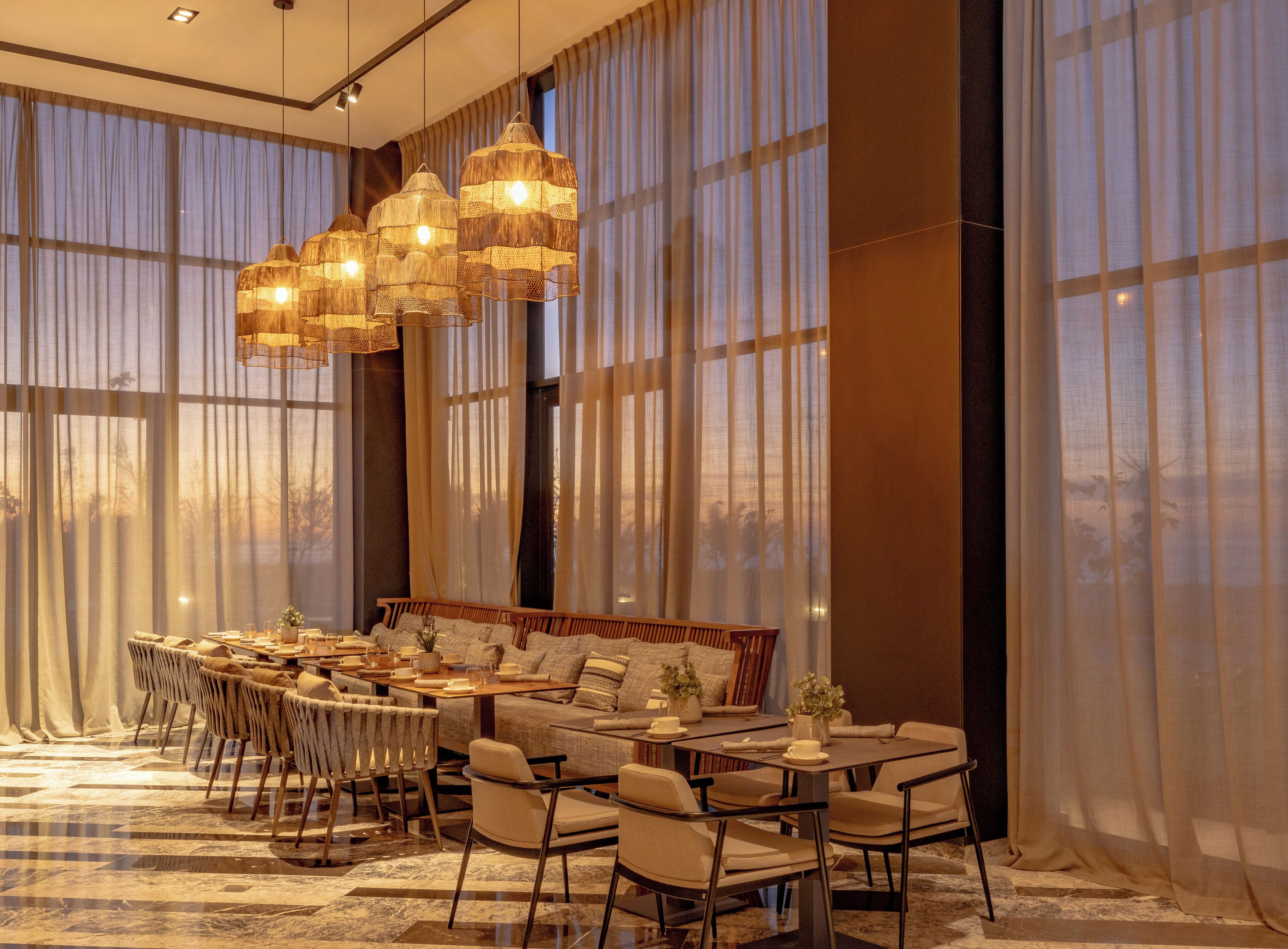 A modern dining area with a round table, several chairs, a sofa, and a large window with sheer curtains.