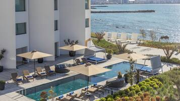 A rooftop pool area with lounge chairs and umbrellas, overlooking a body of water and a city skyline.