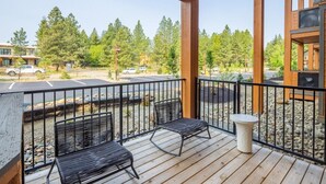 A wooden deck with two metal chairs and a table, overlooking a parking lot and trees.