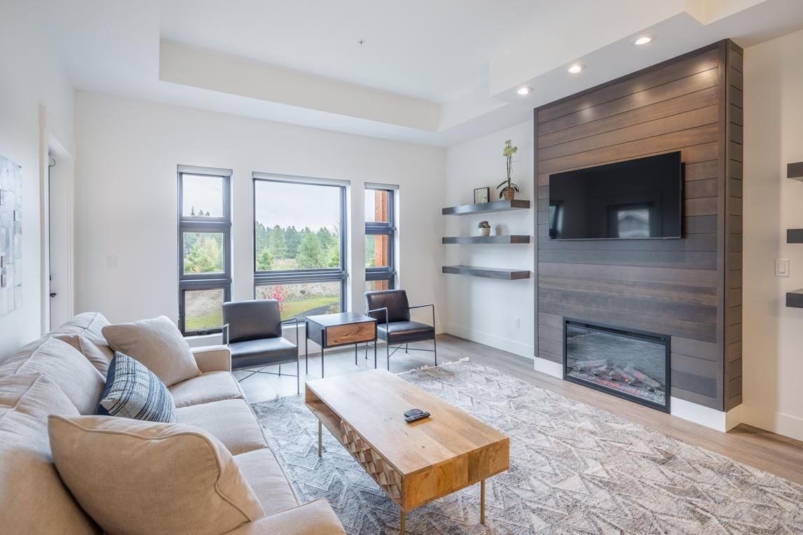 A modern living room with a sofa, coffee table, and chairs. There is a fireplace, a TV mounted on the wall, and large windows offering a view of the outdoors.