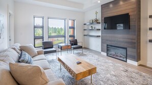 Un salon moderne avec un canapé, une table basse et des fauteuils. On y trouve une cheminée, un téléviseur fixé au mur et de grandes fenêtres qui offrent une vue sur l’extérieur.