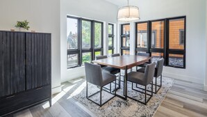 A dining area with a wooden table and six chairs, large windows with black frames, a hanging light fixture, and a dark wooden cabinet. - Golf View Condo & Suncadia Pool Access (Cle Elum)