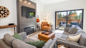 A modern living room with a fireplace, a large TV mounted on the wall, and a view of the outdoor deck through large sliding glass doors. - Golf View Condo & Suncadia Pool Access (Cle Elum)