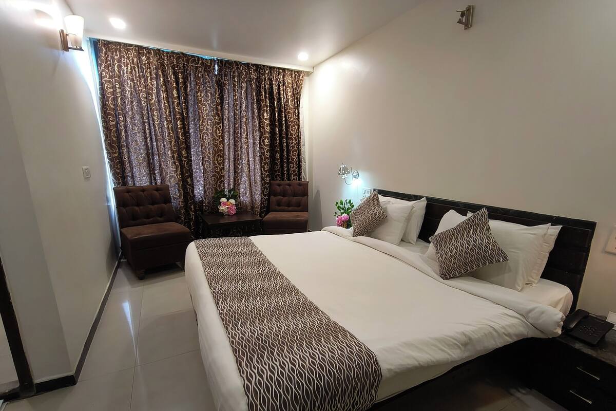 A modern bedroom with a large bed, two brown armchairs, a small table with a vase of flowers, and patterned curtains.