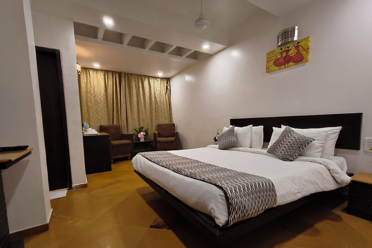 A hotel room with a large bed, a desk with a chair, a small table with a vase, and a painting on the wall.