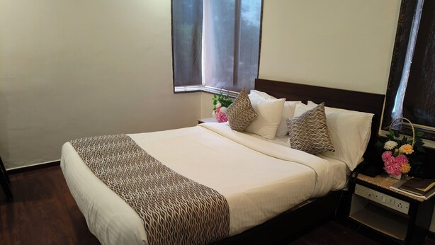 A neatly made bed with patterned bedding, a bedside table with a vase of flowers, and a window with blinds. - Hotel Heritage 2001-Nagpur (Nagpur)