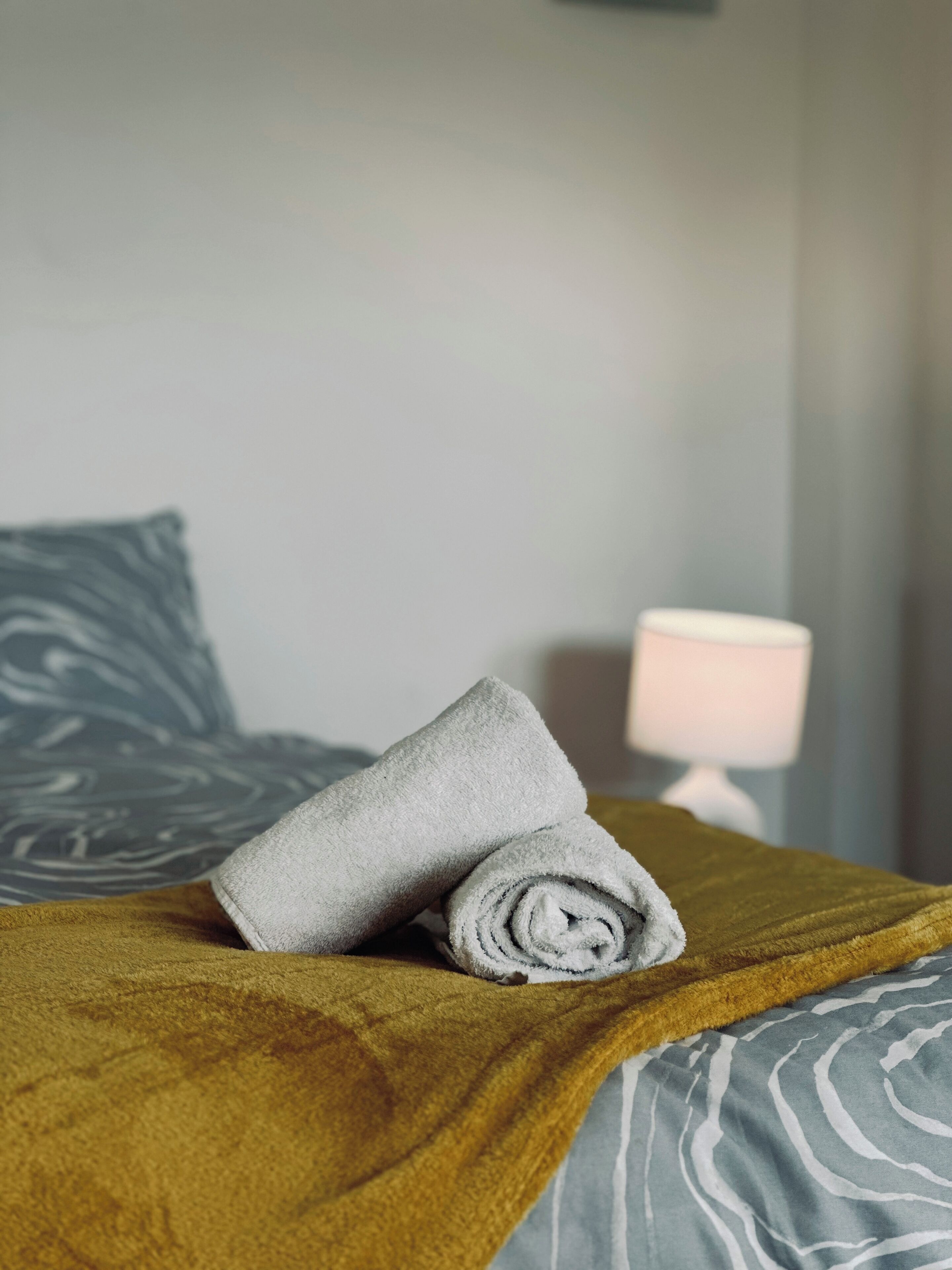 A neatly made bed with a rolled-up towel, a patterned blanket, and a lamp in the background.