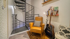 A staircase with a metal railing, a yellow armchair with a patterned pillow, a guitar, and a shelf with books and decorative items. - Maison Bossanne (Barbières)