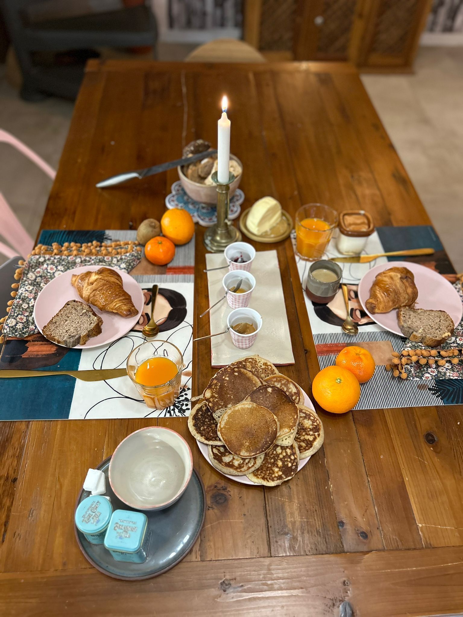 A wooden dining table set with plates of food, including bread, pancakes, and oranges. There is a candle in the center, and various condiments and drinks are also visible.