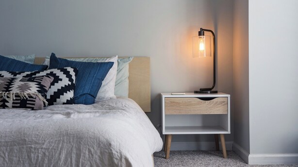 Un lit bien fait, avec une couette blanche et des coussins à motifs, une table de chevet avec une lampe, et un mur de couleur claire.