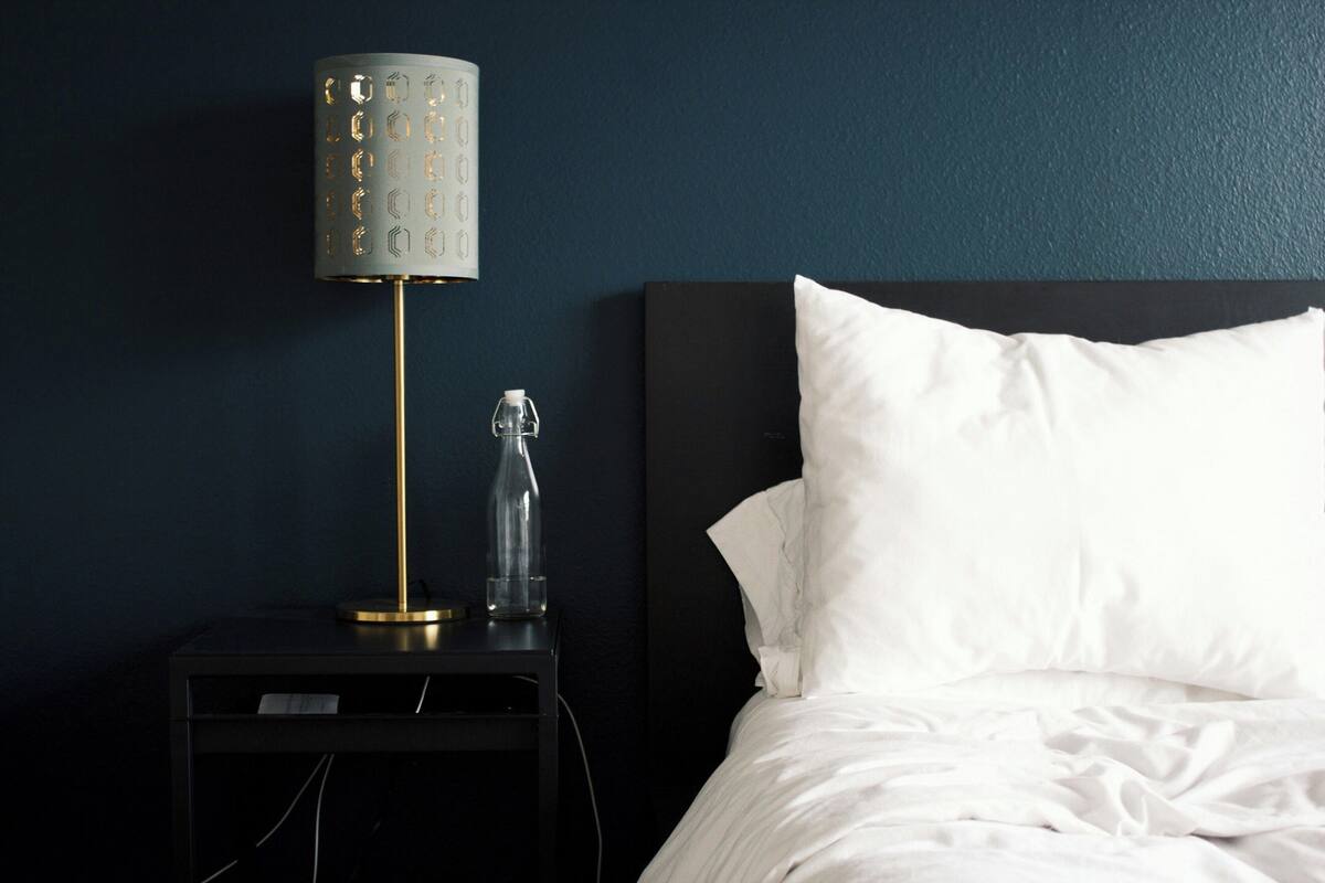 A neatly made bed with white bedding, a black nightstand with a lamp, and a clear glass bottle.