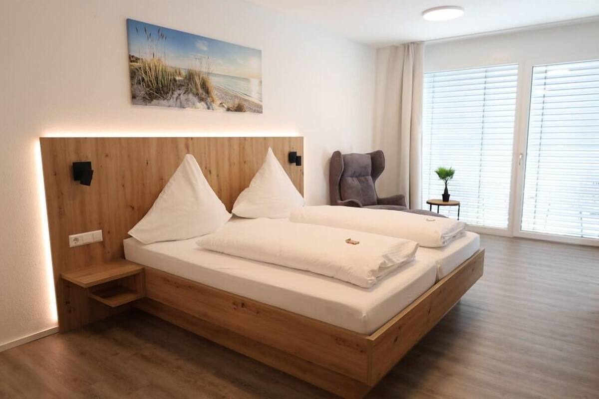 A modern bedroom with a wooden bed frame, two pillows, a nightstand, a chair, and a small table with a plant.