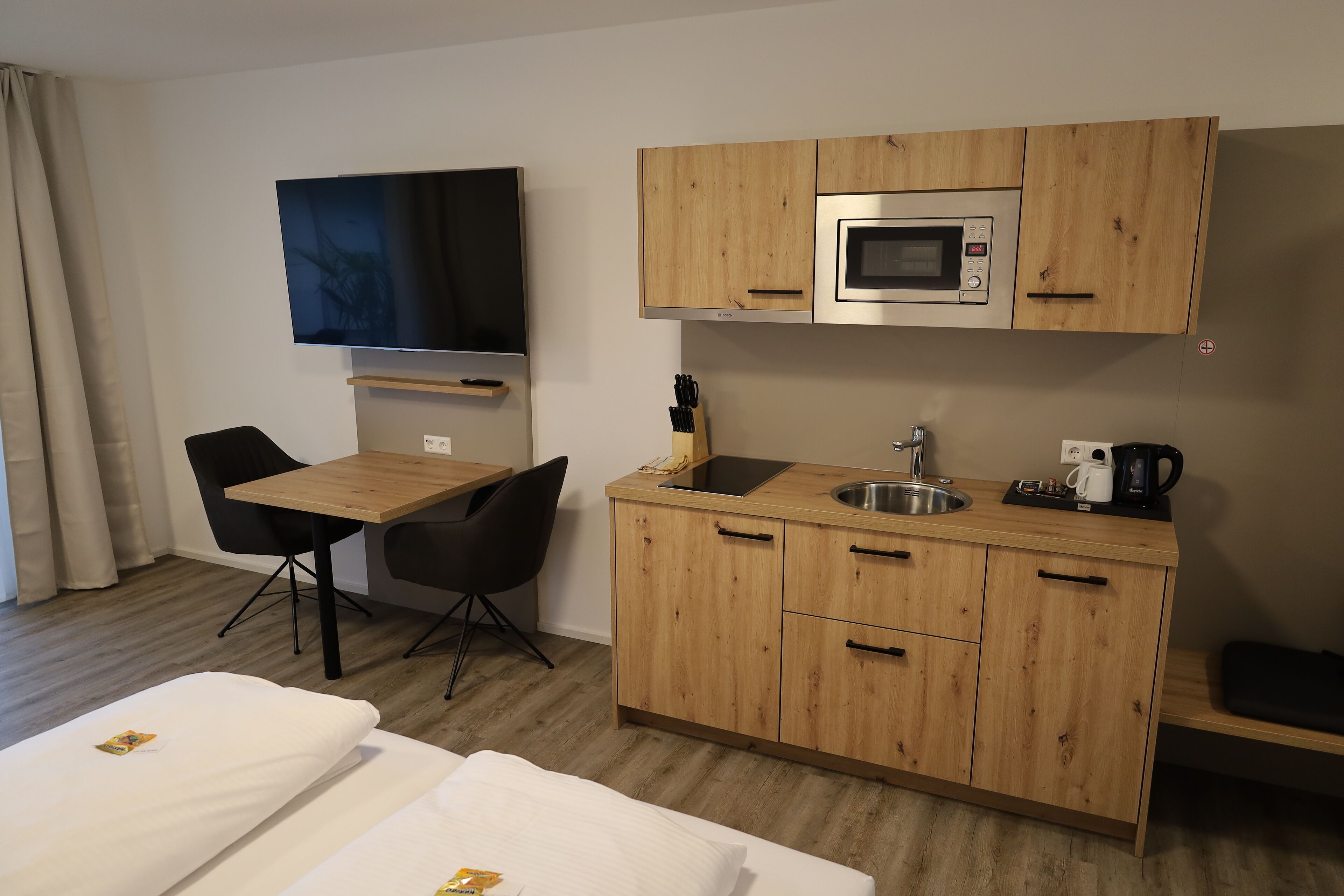 A compact hotel room with a kitchenette including a microwave, sink, and coffee maker. There is a TV mounted on the wall and a small dining table with two chairs.