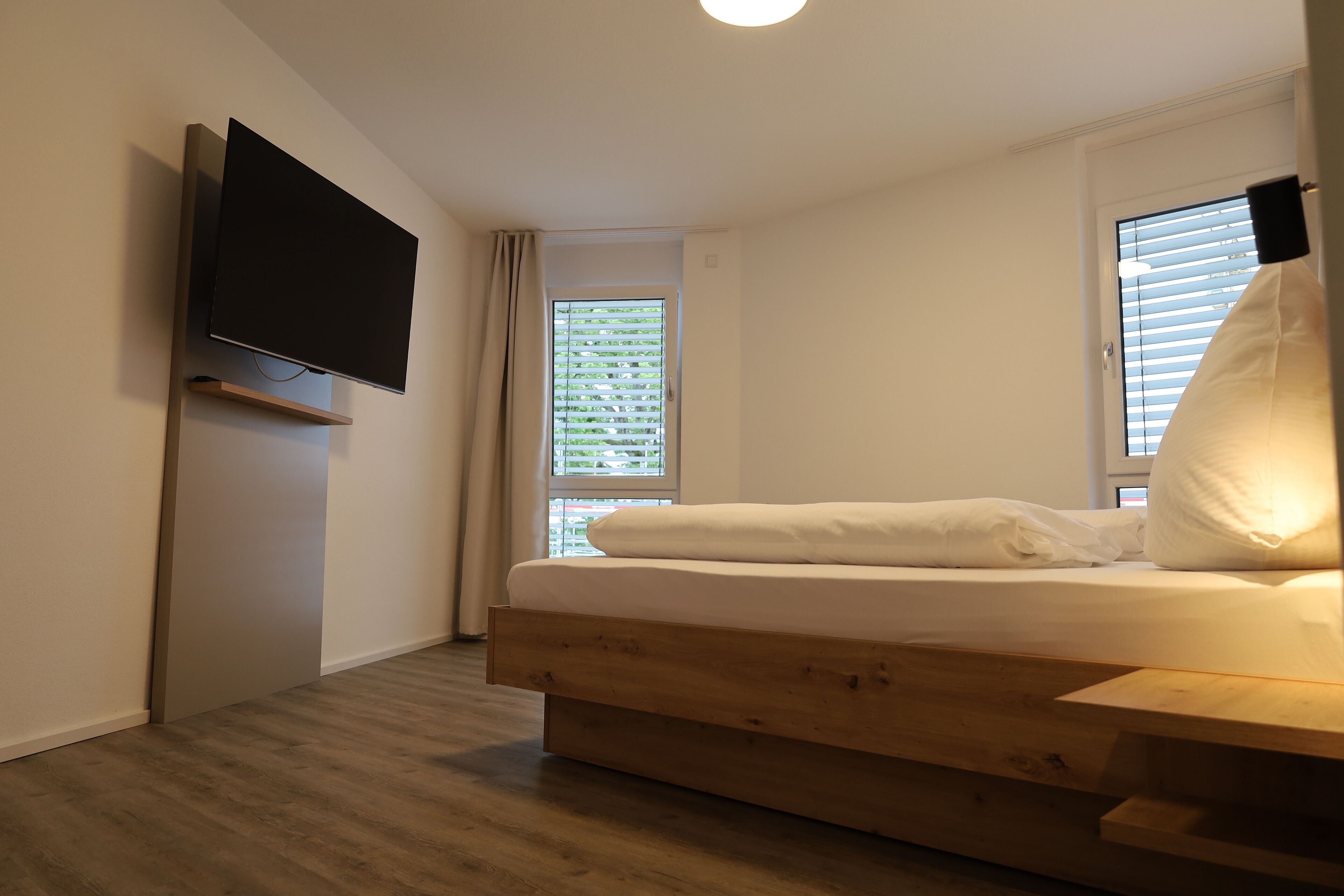Quarto moderno com cama de madeira, televisão montada na parede e janela com persianas.