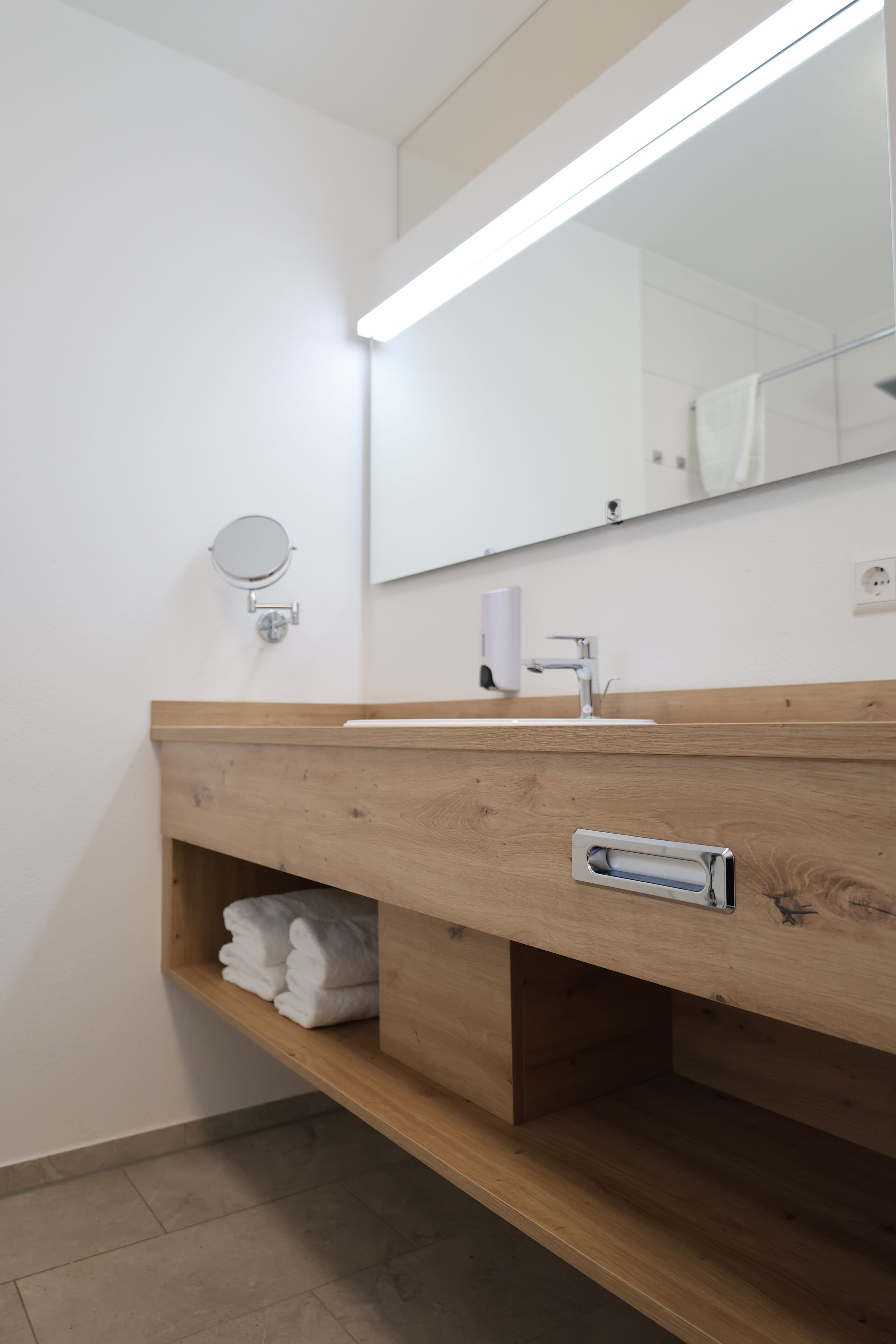 A modern bathroom with a wooden vanity, a round mirror, a towel rack, and a hand dryer.