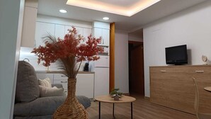 A modern living room with a sofa, a coffee table, a vase with red foliage, a television, and a kitchen in the background.