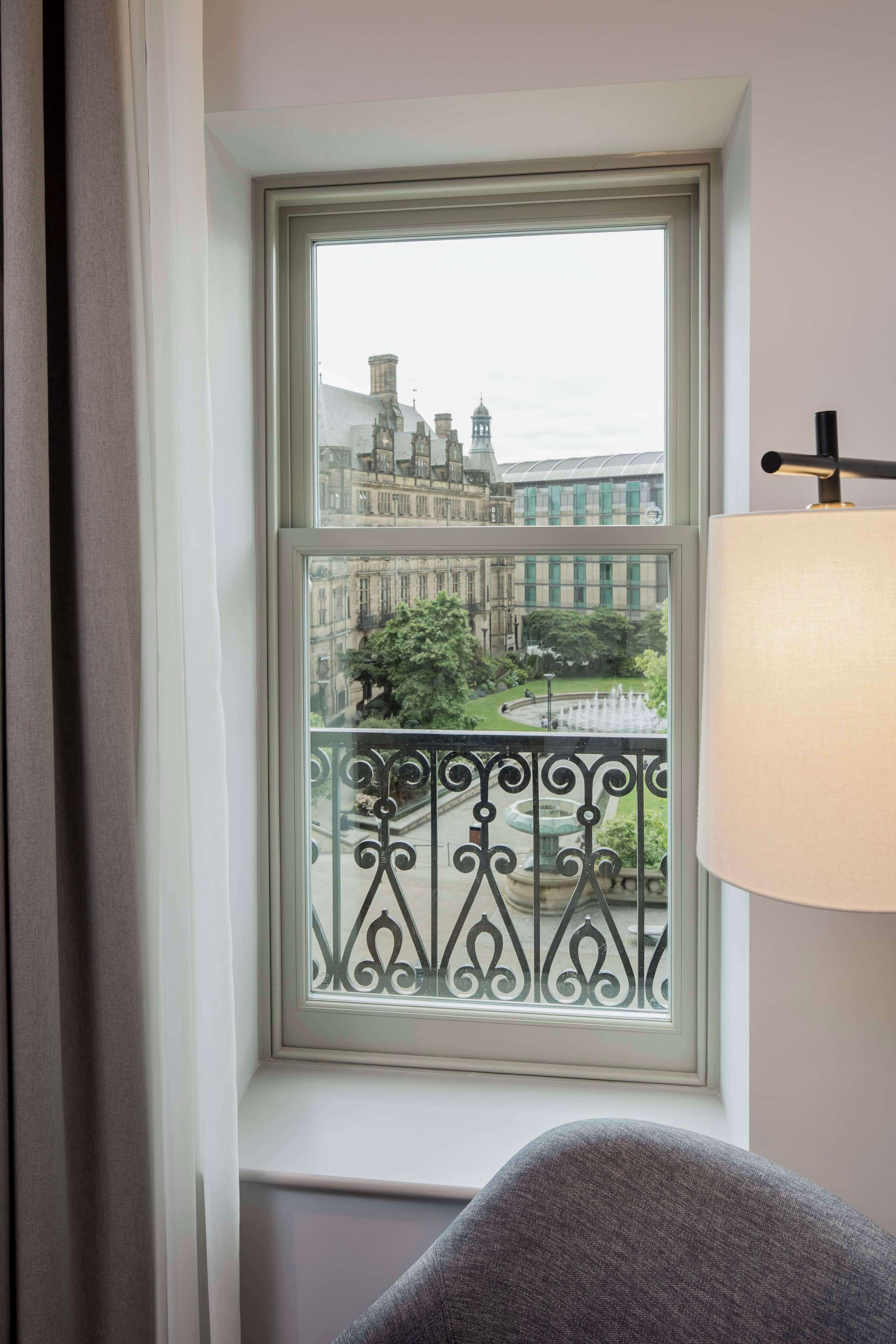 Una habitación con ventana que da a un paisaje urbano, un balcón con barandilla de hierro ornamentada y una lámpara a la derecha.