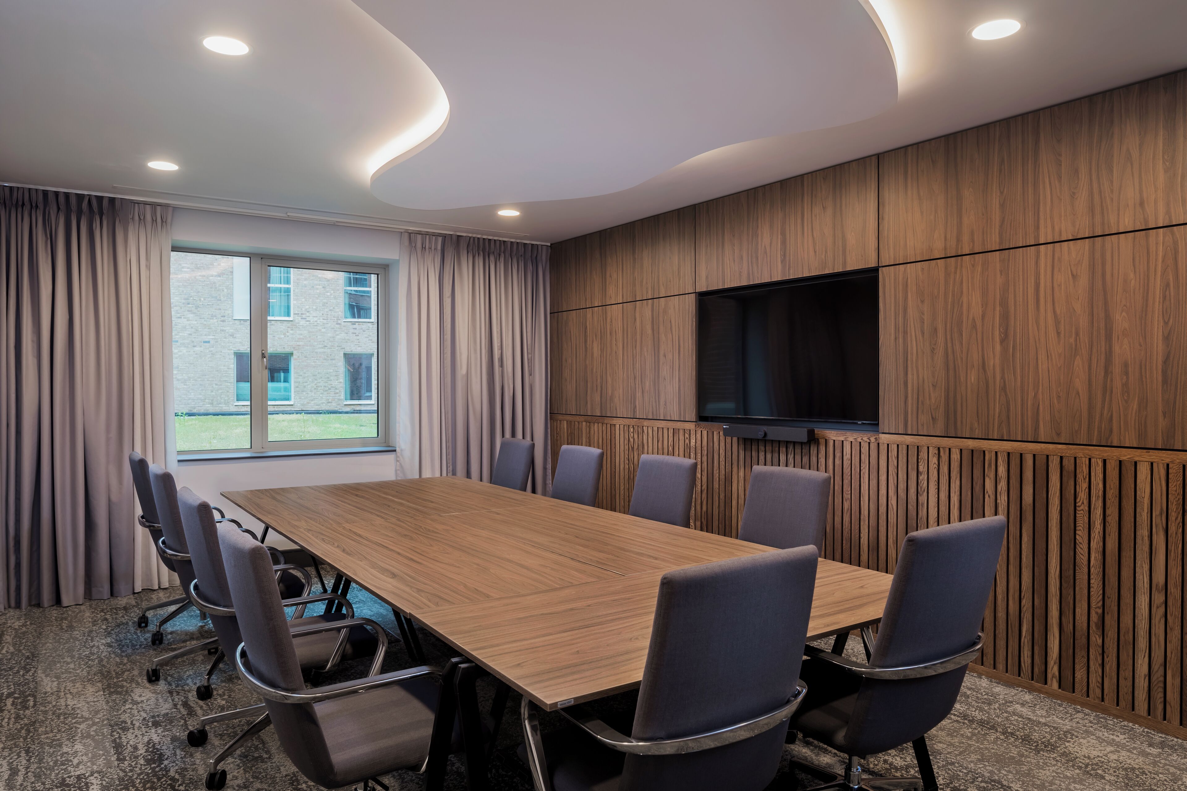 A conference room with a long table, six chairs, a flat-screen TV, and a window with curtains.